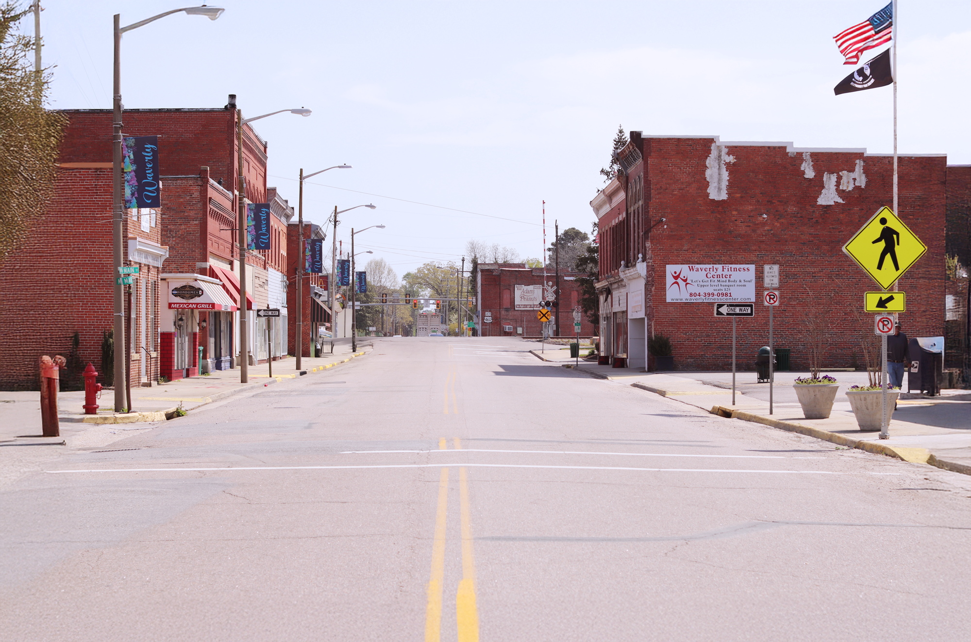 A quiet, empty street in downtown Waverly, Virginia, lined with old red-brick buildings on both sides. The street features storefronts including a diner with a red and white striped awning and a fitness center. American and POW/MIA flags fly on a pole to the right. A pedestrian crossing sign and railroad crossing sign are visible in the distance, along with flower planters and vintage streetlamps. The overall atmosphere is calm and small-town. This is the home of the first office of the improvement association.