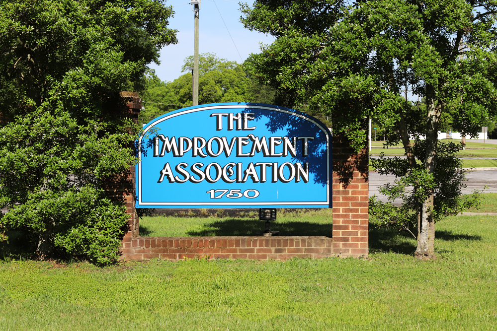 The Improvement Association sign is framed by bricks, with two seven-foot trees on both ends, in a grassy area. sign reads The Improvement Association with the building number 1750 at the bottom.